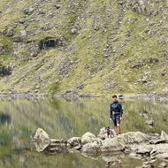 Looking across a mountain lake at the bottom of a huge mountain. A tiny dog and person in the background