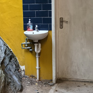 On the right of the image, a metal urinal in a men's toilet. In the centre the door and a sink. On the left hand side, an enormous rock formation with vines/leaves/moss which forms the left-hand wall.