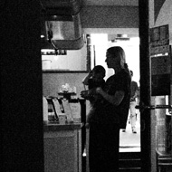 A woman holding her baby silhouetted in a coffee shop. Lit from behind, the natural shapes her body makes contrast against the straight lines and square angles of the doors, windows, and wall.