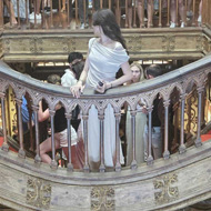 On the top floor of the bookshop, in the background are bookshelves, above the bookshelves are art deco style paintings decorating the walls. The majority of the room is taken up by the wooden frame of a grand staircase, in the centre is a bridge which leads to the steps. Through the wooden frame, you can see downstairs where the bottom of the red steps are visible along with more bookshelves. Both floors are full of people admiring the beauty and taking photos.