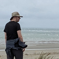 Two people and a dog make their way up a bank of sand dunes and sea grass towards the top beyond which the sea is visible in the back ground of the picture