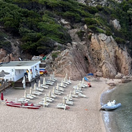 A scenic view of a small sandy beach nestled in a rocky cove with clear blue water. People are walking along the beach and swimming. Steep, rugged cliffs covered in greenery rise behind the beach, and a few beach chairs and umbrellas are visible on the left side near a white structure.