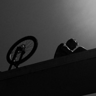 A photo taken from below Destructor Bridge's single arc-shaped solid support. A man is sitting on top of the support entirely in shadow except for a thin outline of light. Behind him is a bicycle. He is about to ride down. Light cascades from the top-right of the frame towards his face, and shadows on the bridge supports below frame him in the center of the image.