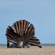 A wide pebble beach below a wide blue sky. On the beach a large sculpture of a scallop shell made from curving sheets of metal is set against the sky