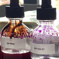 A line of labelled bottle droppers containing coloured inks, behind a piece of paper with drops of each colour.
