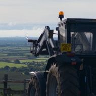 A dark coloured tractor with a bailing fork on the front faces towards a wooded valley with low, bright blue-white clouds in the distance