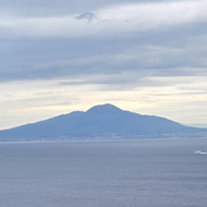 A stretch of water with Mount Vesuvius on the other side. Clouds above look a bit like it is coming from the volcano. In the foreground is green shrubbery framing Vesuvius