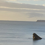 Glorious blue sea meets blue sky with a few whispy clouds. In foreground a tennis court and green trees are enjoying being lit by the sunlight.