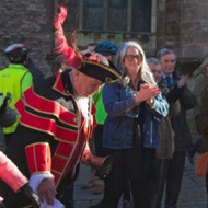 A crowd has gathered for the official welcoming of a new hare statue. The town crier resplendent in his red uniform bows to the artist. The hare in the right foreground is decorated with iconic scenes of both old and new Cirencester