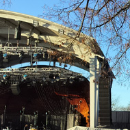 An outdoor stage before the crowd arrive. There's snow on the ground, a large soundsystem, screen and, on the far right, a windmill.