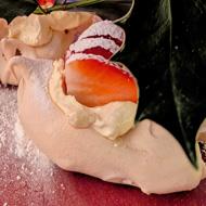 A plate of meringues, cream and strawberries with a sprig of holly on top, against a backdrop of a Christmas tree all lit up