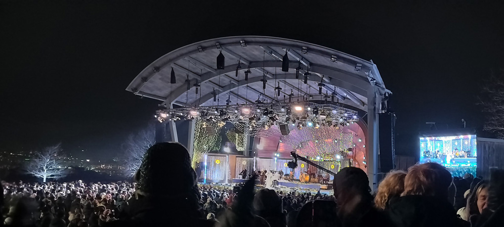 An outdoor stage at night with a large crowd in attendance with a large soundsystem, screen and musicians on stage.
