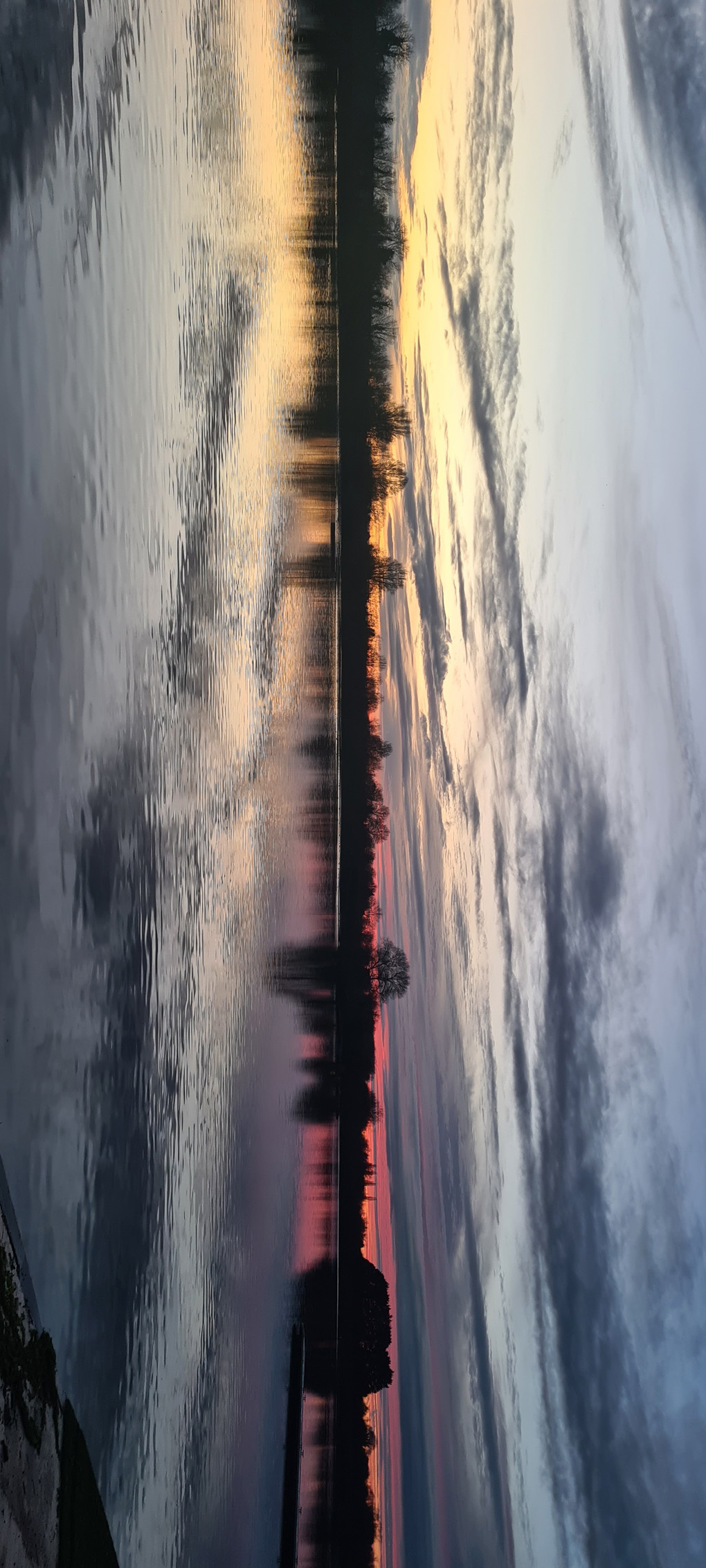 Lake 12 at South Cerney at dusk with yellow and pink sunset reflected in the water. The image is inverted 90 degrees, bringing attention to the light splitting down the centre of the photograph creating a more abstract void-like image.