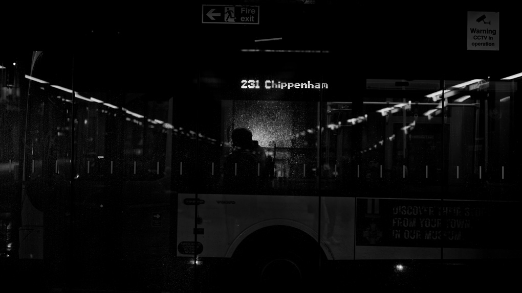 A bus to Chippenham pulling out of bath bus depot, at night, shot from inside the depot. one passenger is visible through the station and bus windows in the center of the frame. the reflection of the station strip lights form a shallow v converging on their head, reminiscent of kubrick's stargate sequence in 2001. raindrops and other glass elements reflect light