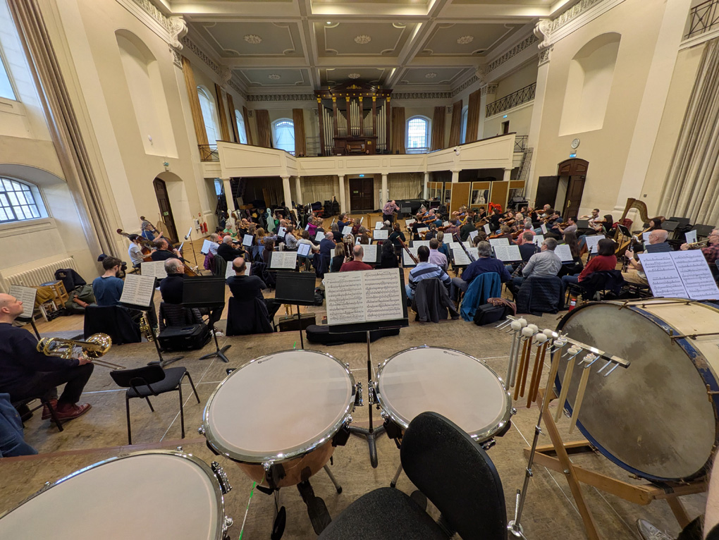 A view from the back of an orchestra, timpani in the foreground, and the orchestra spread out in a large hall.