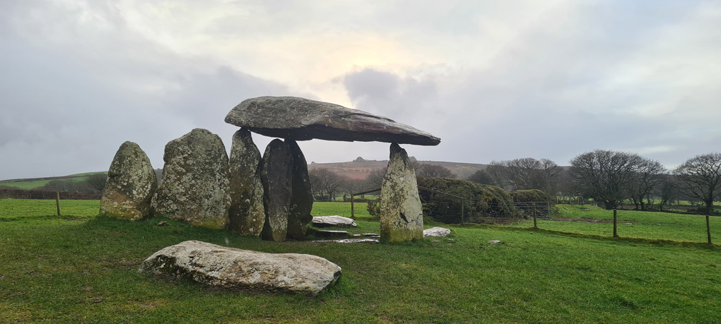 Ancient standing stone monument in green fields. A large flat stone lies upon three pointed standing stones in what appears to be impossible balance.