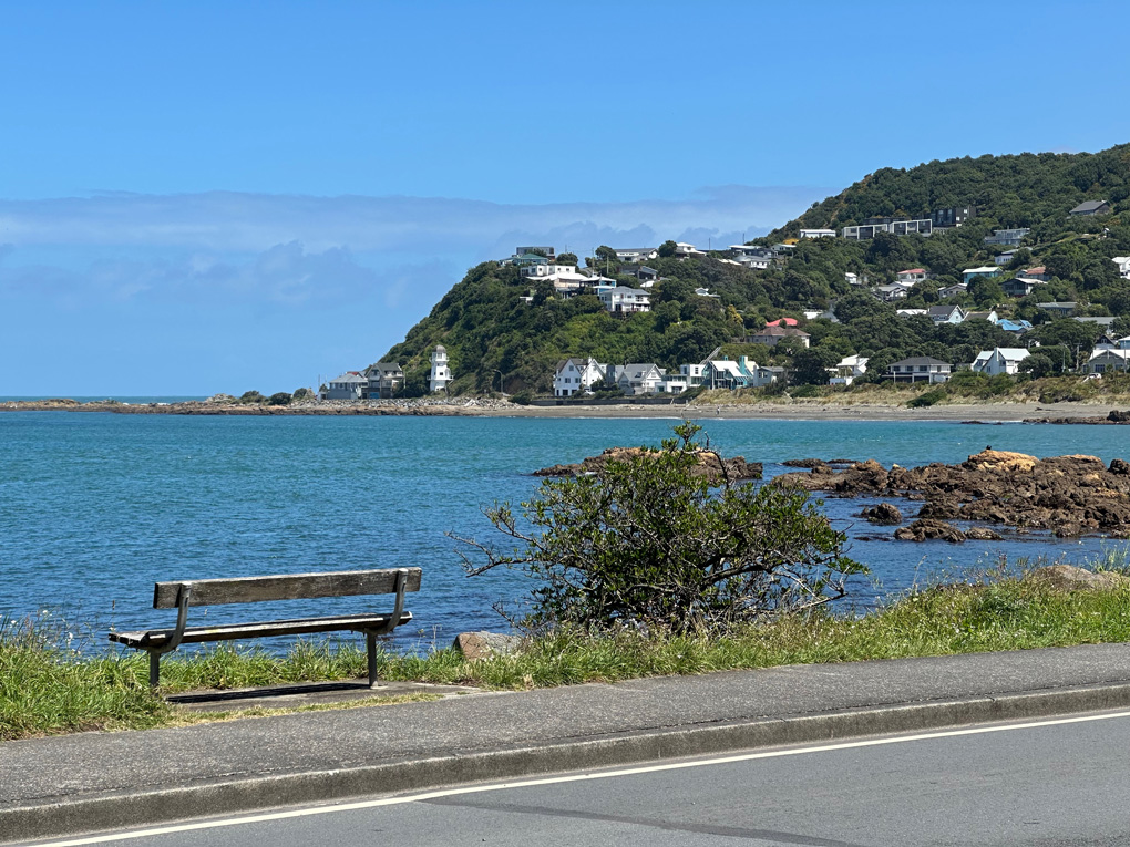 View from Island Bay, Wellington. Looking out over the sea as the coast curves around. Bench in foreground. Hills and lighthouse in distance.