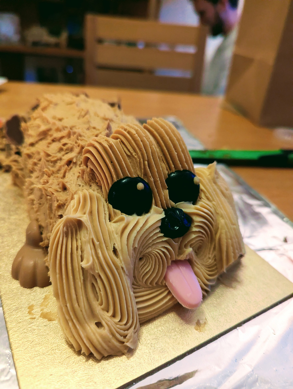 A long cake decorated with lots of swirled icing to look like a fluffy dog. Two jet black eyes and a tongue poke out from the face in the foreground