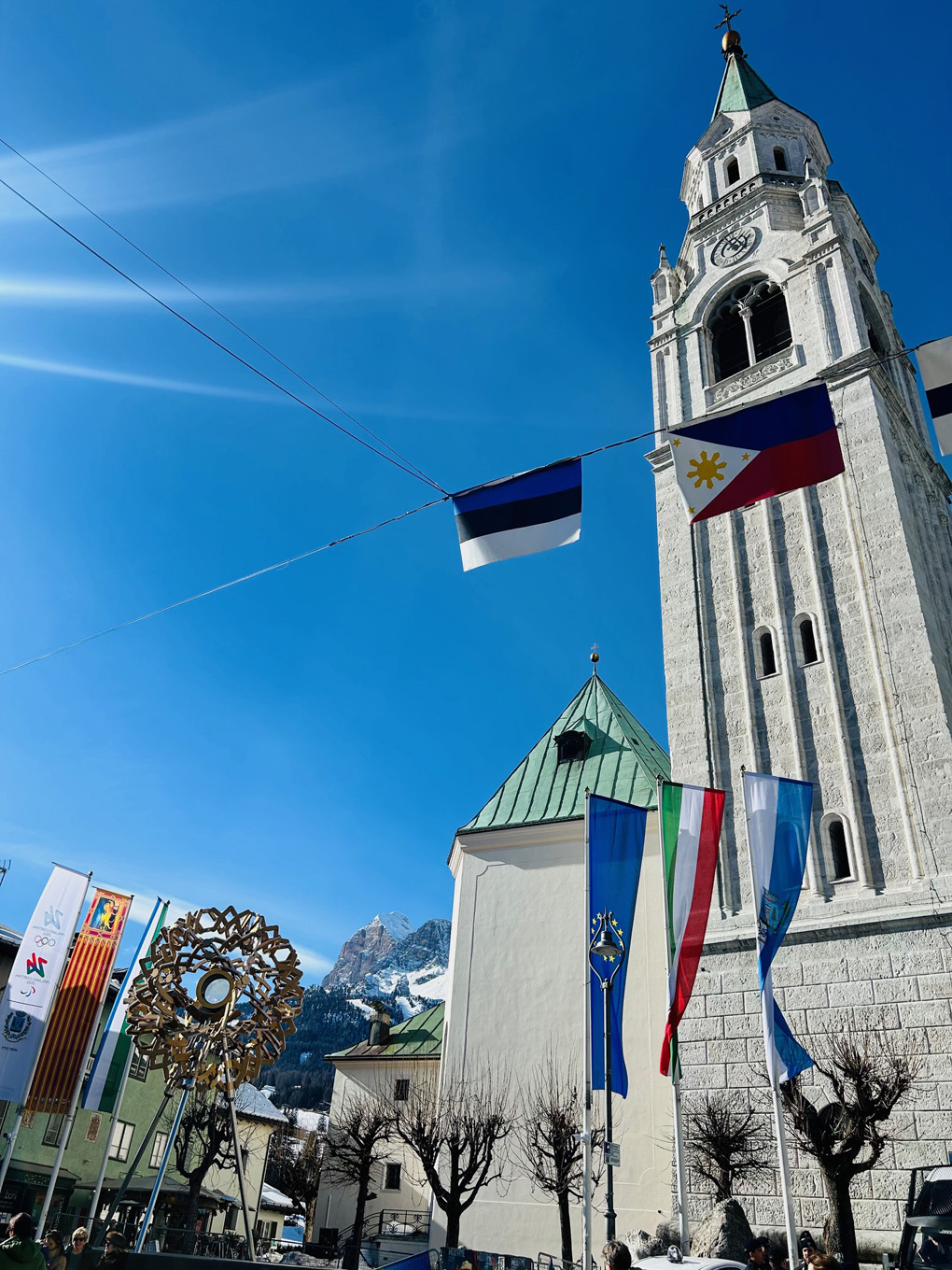 A blue sky, early morning, frames a picture of a large church spire beside which the metal fluted ring of the Winter Olympics cauldron - with its eternal flame - burns. Shot on a town square in the mountain town of Cortina, Italy.