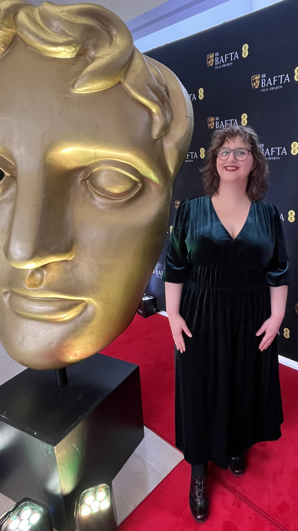 Me, a short brunette woman with glasses wearing a velvet green dress, standing on a red carpet next to a large statue of a BAFTA award