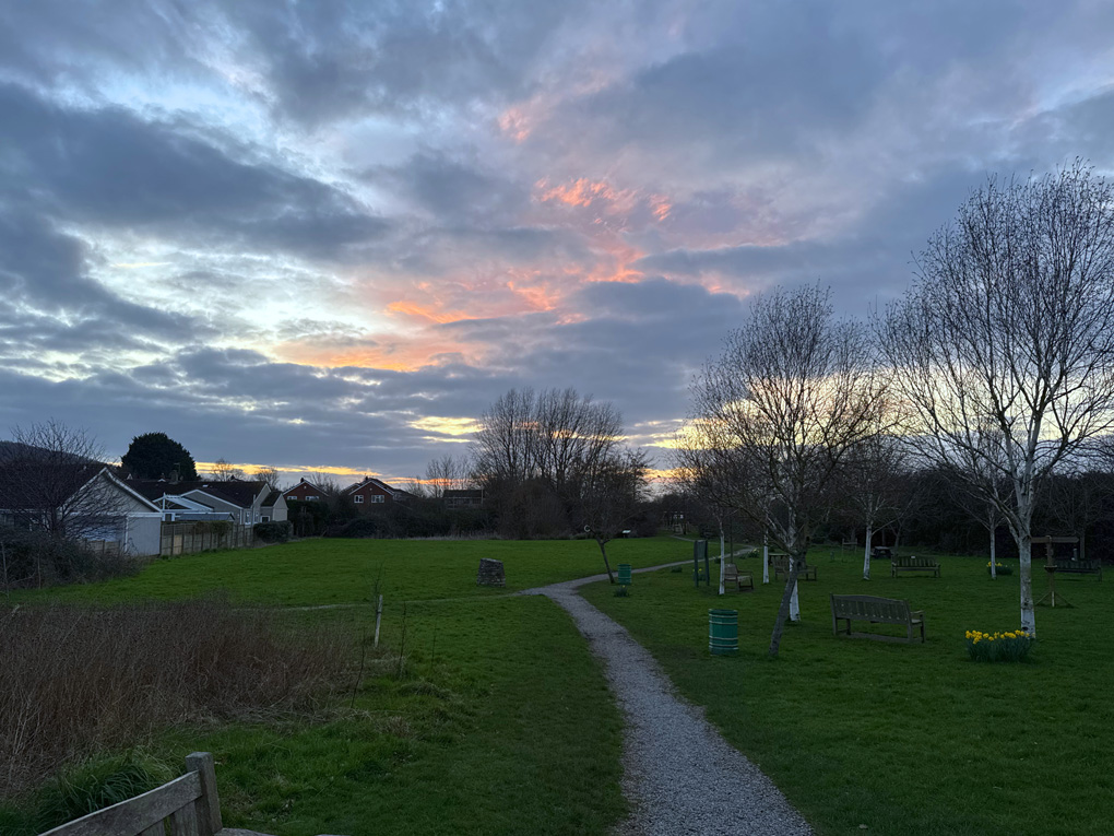 A cloudy sky obscures a deep orange and purple sunset above a green park