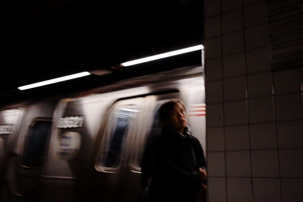A slightly blurred photo of a woman walking along a subway platform in new york with the train moving behind her