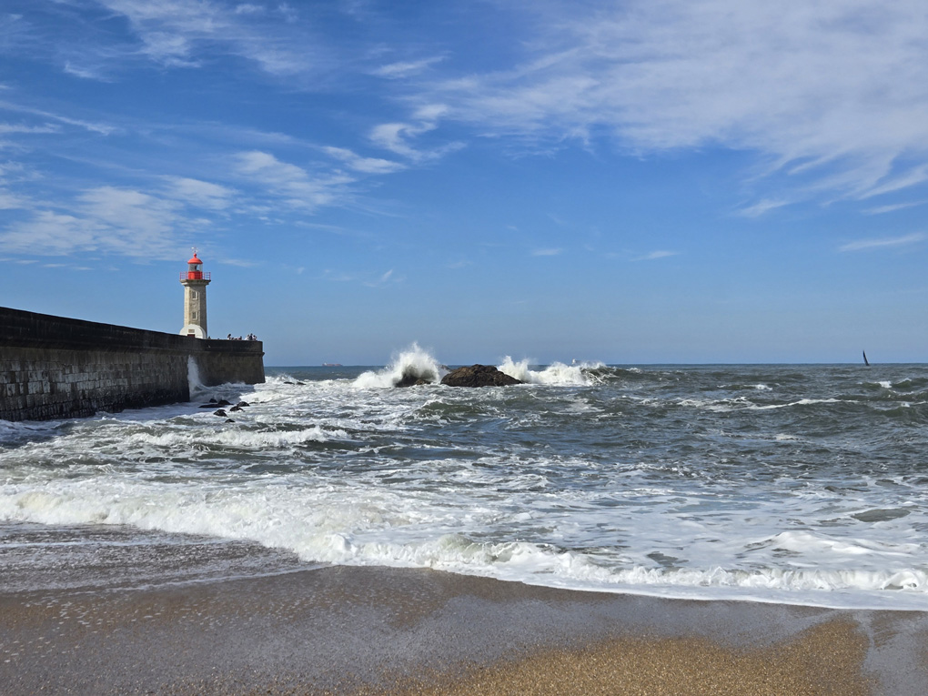 Photo taken from a shoreline. A lighthouse stands on a stone pier. A wave has crashed into a rock a few metres away, sending spray into the air.