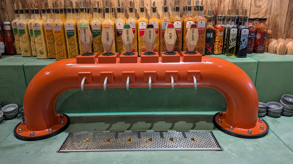 A scene from Nando's. A shelf of the normal sauces available, as normal - however in front of those is a dispenser, looking like the taps in a pub, with a nozzle for each flavour of source. It is a bright orange set of pumps dispensing Nando's sauce on-tap in flavours (Left to Right) - Lemon & Herb, Medium, Wild Herb, Garlic, Hot, Extra Hot. There is a drip tray under the whole thing.