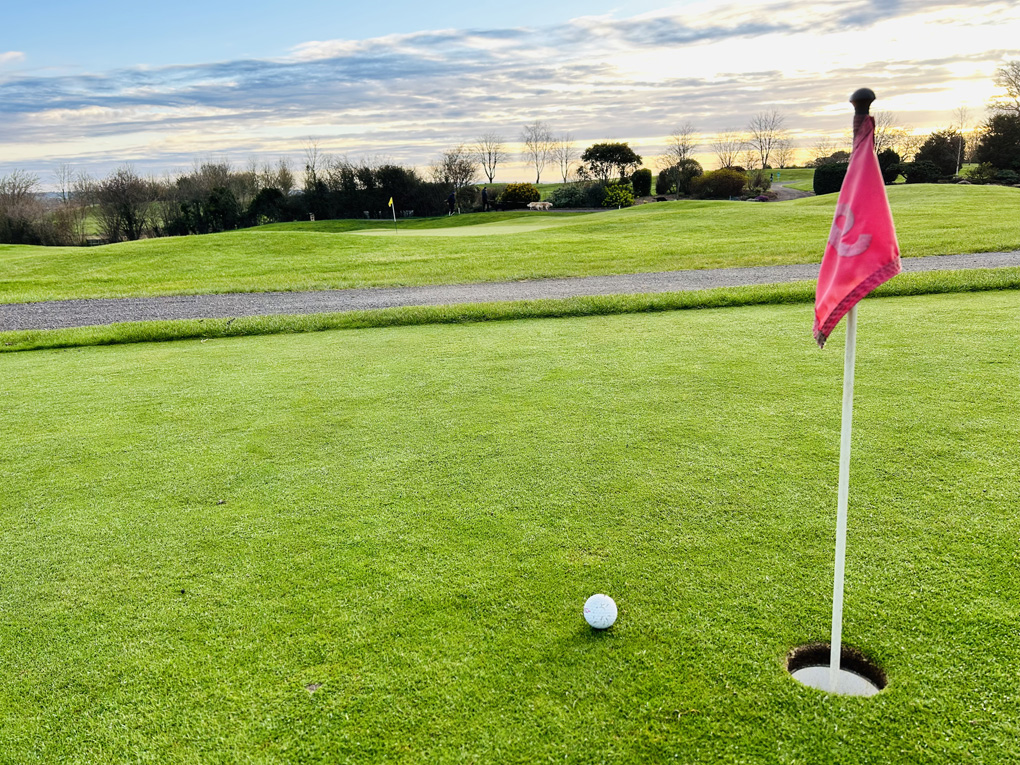 Sunrise at a golf club. A golf ball site beside a hole with the flag in, and in the background the sun rises and dapples the clouds in a mixture of red and orange.