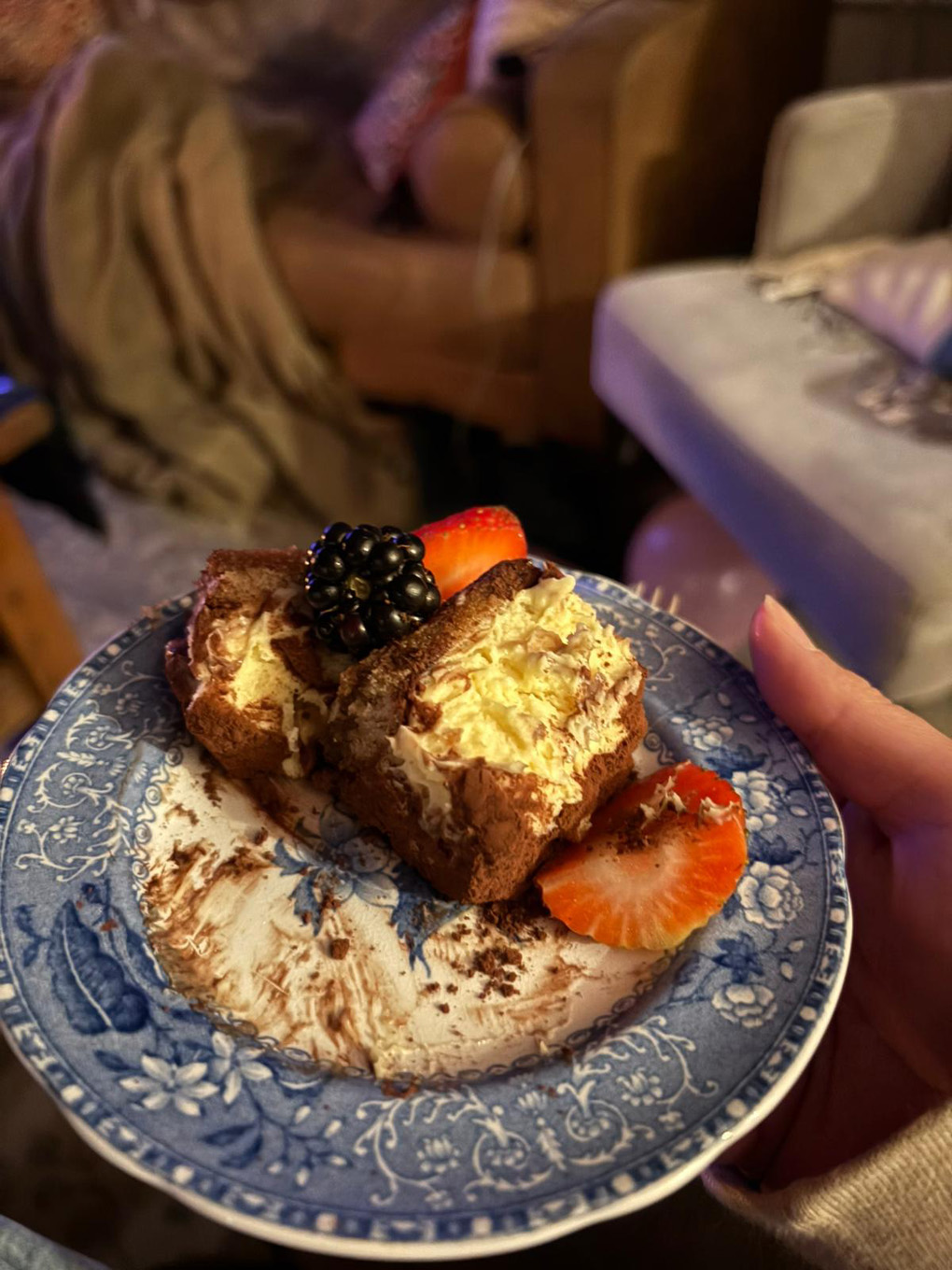 A slice of tiramisu lays on a blue and white plate with two slices of strawberry and a blackberry on top