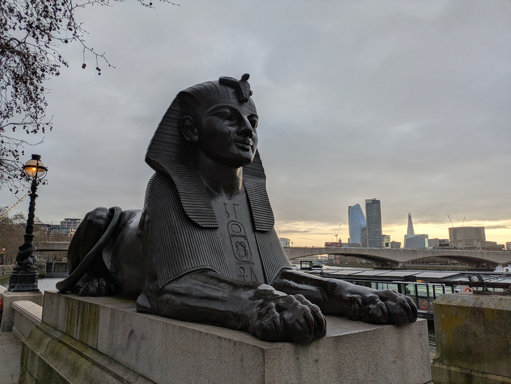 A large dark bronze sphinx on a plinth with the river Thames behind and high rise city buildings in the distance on a slightly grey early morning with the sun just starting to glow through on the horizon