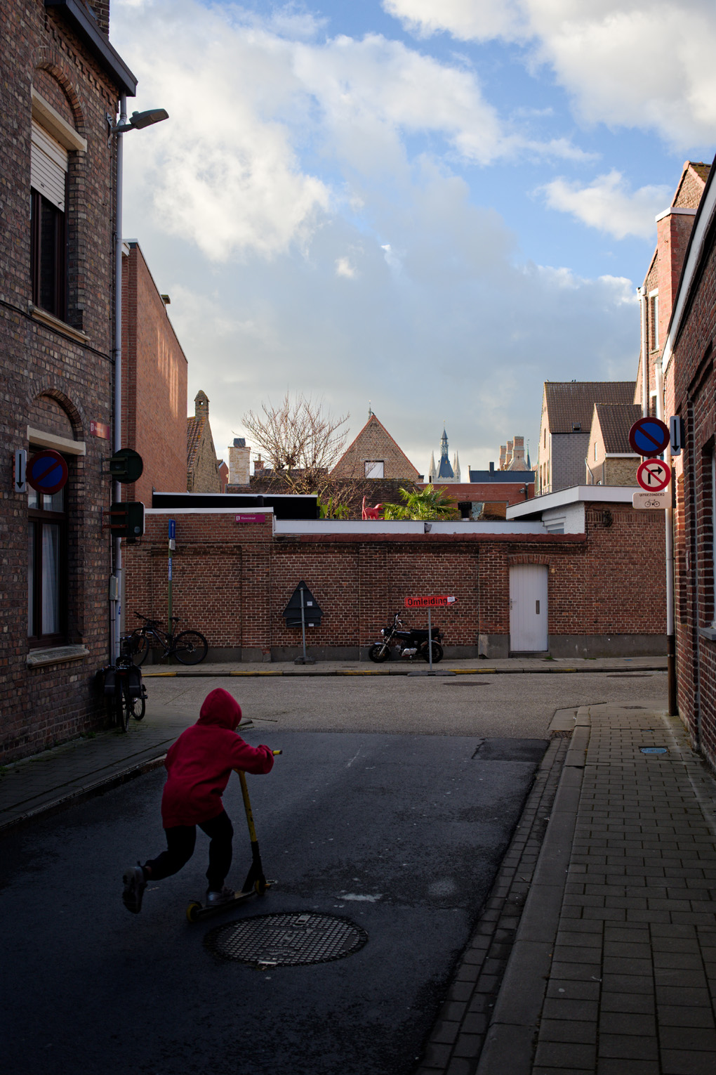 On a small side street in Ieper, a child in a red hoodie rides off on his scooter past the red brick walls and under a bright blue sky