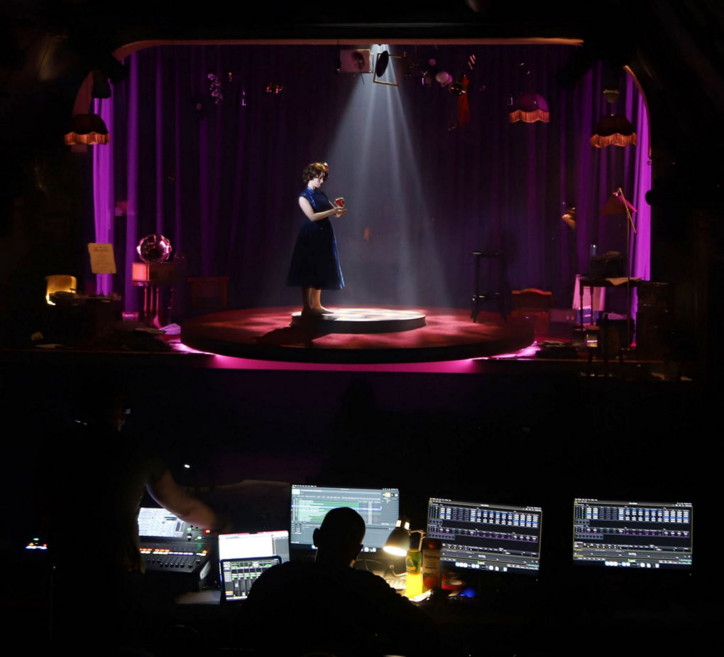The main subject is a single figure on spotlighted on stage. In the foreground are technicians silhouetted by the lights from their many monitor screens.