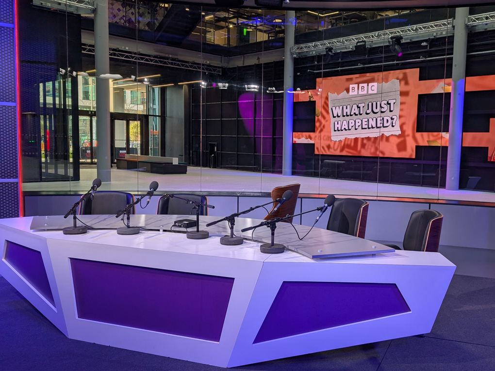 In the foreground, a long, purple and white desk set up for a panel show with five empty chairs and five microphones for the host/panel. The set itself is mainly purple. Behind the desk is a window which looks out over a foyer space, but at the back of the foyer is a large digital display with the show's logo - an orange design and the words 