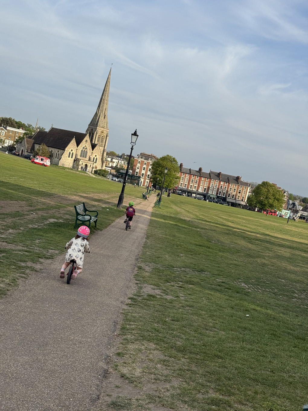 Two children ride away from the camera along a path across Blackheath, wearing helmets, heading toward a Victorian church spire. A park bench and lamp post sit to the left, with London terraces in the background under an evening sky.