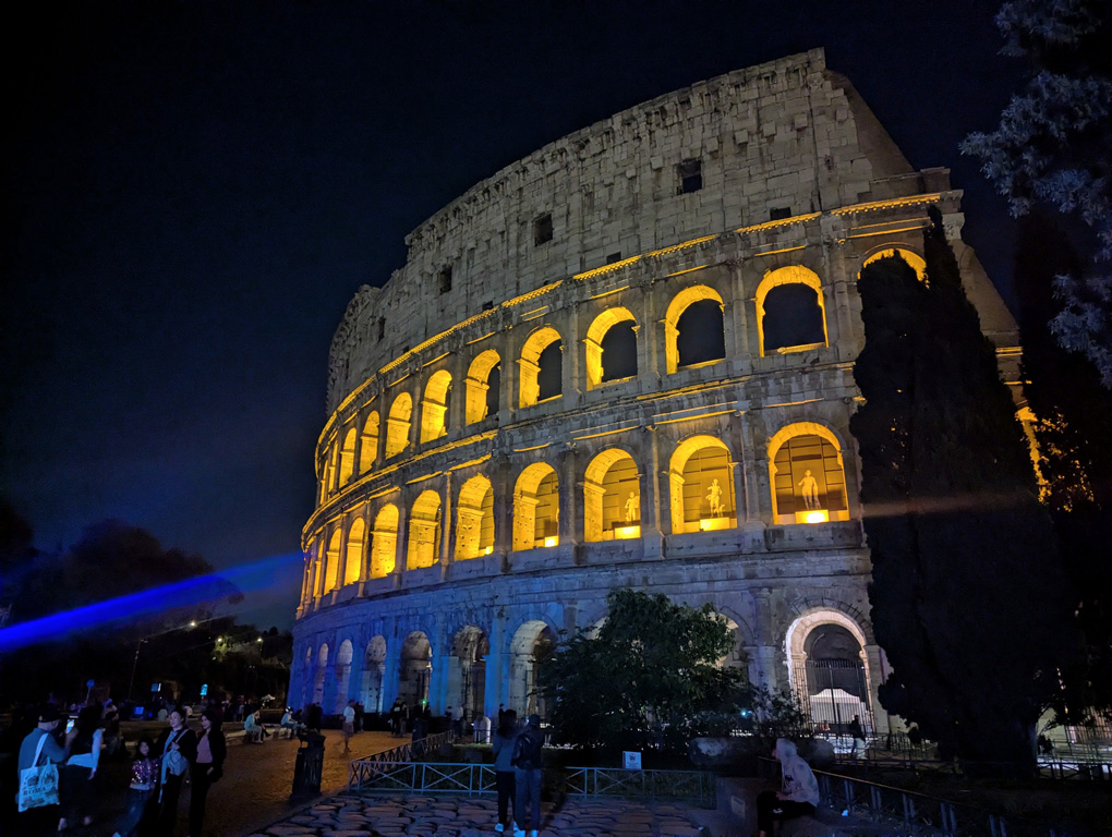 The Rome Colosseum at night, with each recessed arch lit in dark orange light illuminating the statues in each, as they would have been in place in ancient rome
