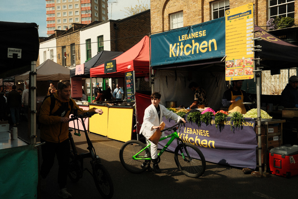 a kid in a white karate gi, on a green bike, is stopped in front of a colorful lebanese food stall in broadway market. The light frames and highlights his face with warmth, contrasting the shadows around him.