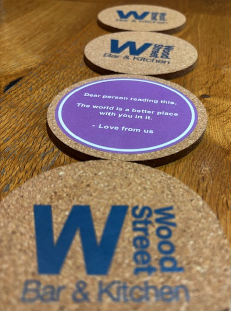 A line of beer mats with one showing the underside that shows the message 'Dear person reading this, the world is a better place with you in it' - Love from us