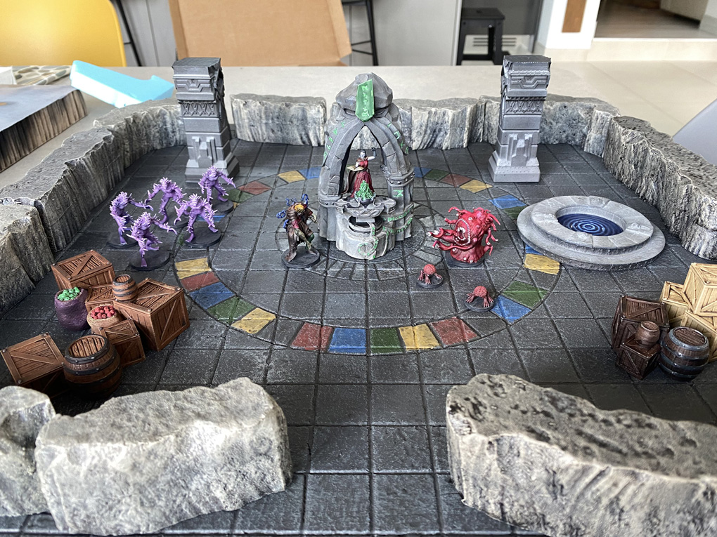 A tabletop RPG dungeon scene featuring painted miniatures on a gridded stone-tile game board enclosed by rocky walls. The setup includes a central stone archway with a robed figure, several purple tentacled creature miniatures, a red multi-eyed monster, a corrupted warrior figure, a swirling well, decorative stone pillars, and scatter terrain including wooden crates and barrels. The board features a circular pattern of coloured tiles in red, blue, green, and yellow.
