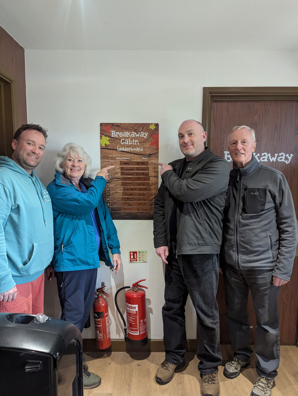 Four family members standing around an escape room leaderboard of escape times, pointing at their team at the top.