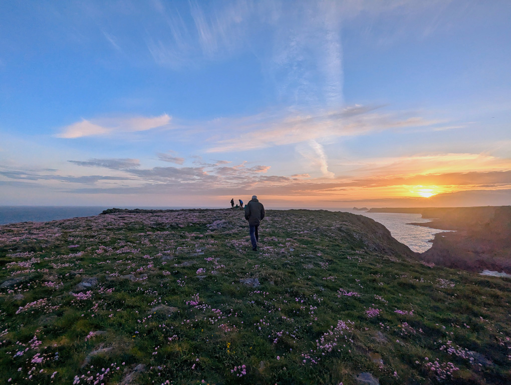 A few figures in silhouette walking along a cliff top with the sea stretching before them and a glowing sunset in the background