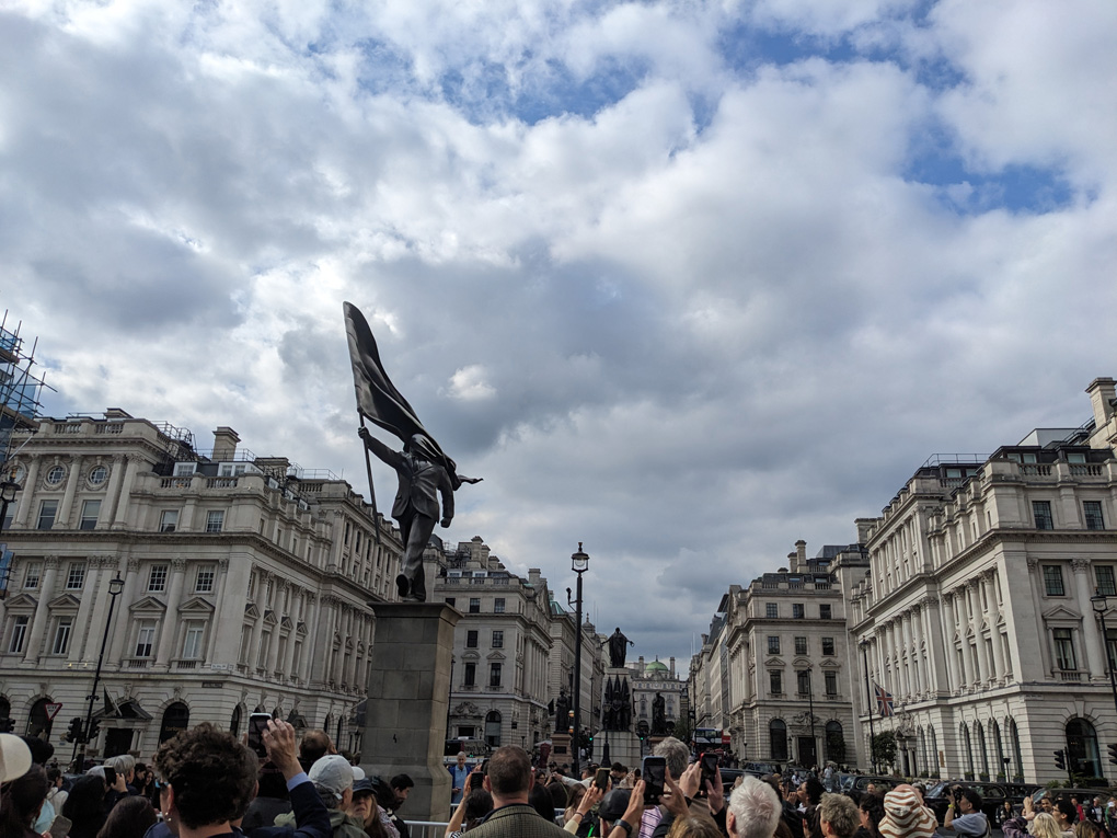 A statue surrounded by important buildings, embassies, government and royalty. One of many statues in the area. The person in this statue is carrying a flag which has blown back to cover their face. They are marching forward off the plinth.