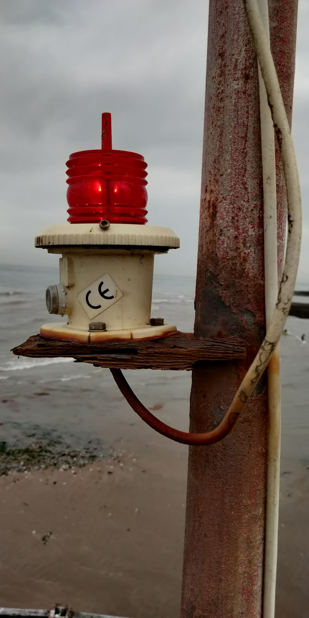 Very rusty bracket suporting a red navigation lamp, with seascape behind.