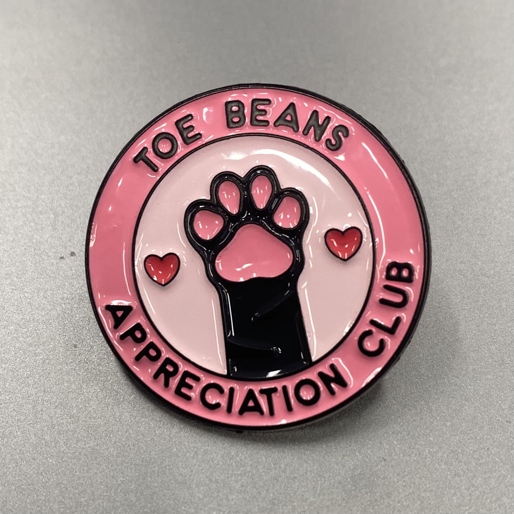 Round pink enamel pin featuring a black cat paw with pink toe beans at the centre, flanked by two red hearts. The text 