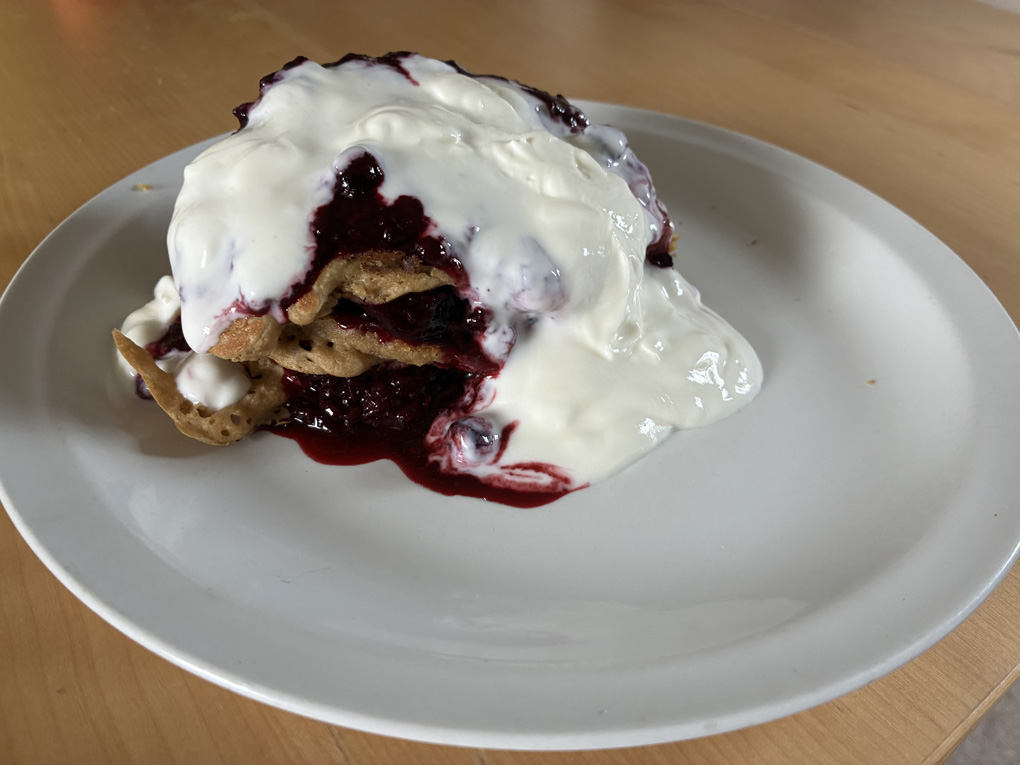 Three small round pancakes stacked on a plate with blue berry compote and white younger oozing from between the layers and all over the top.