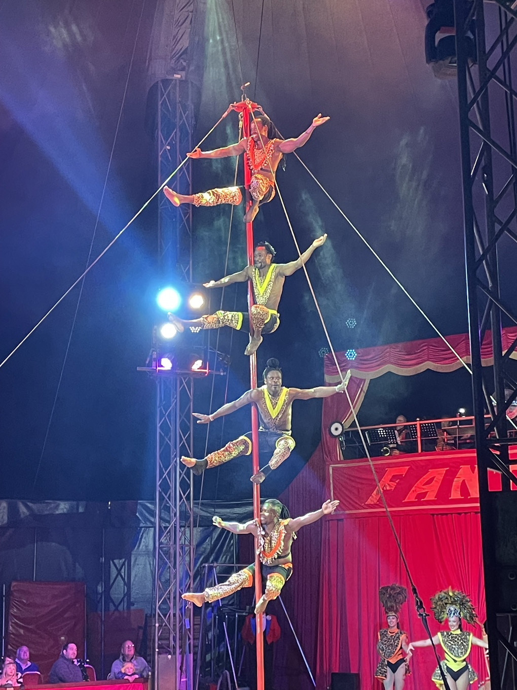 Four brightly dressed circus acrobats balance vertically on a tall red pole, one above the other, with their arms stretched out under colourful stage lights.