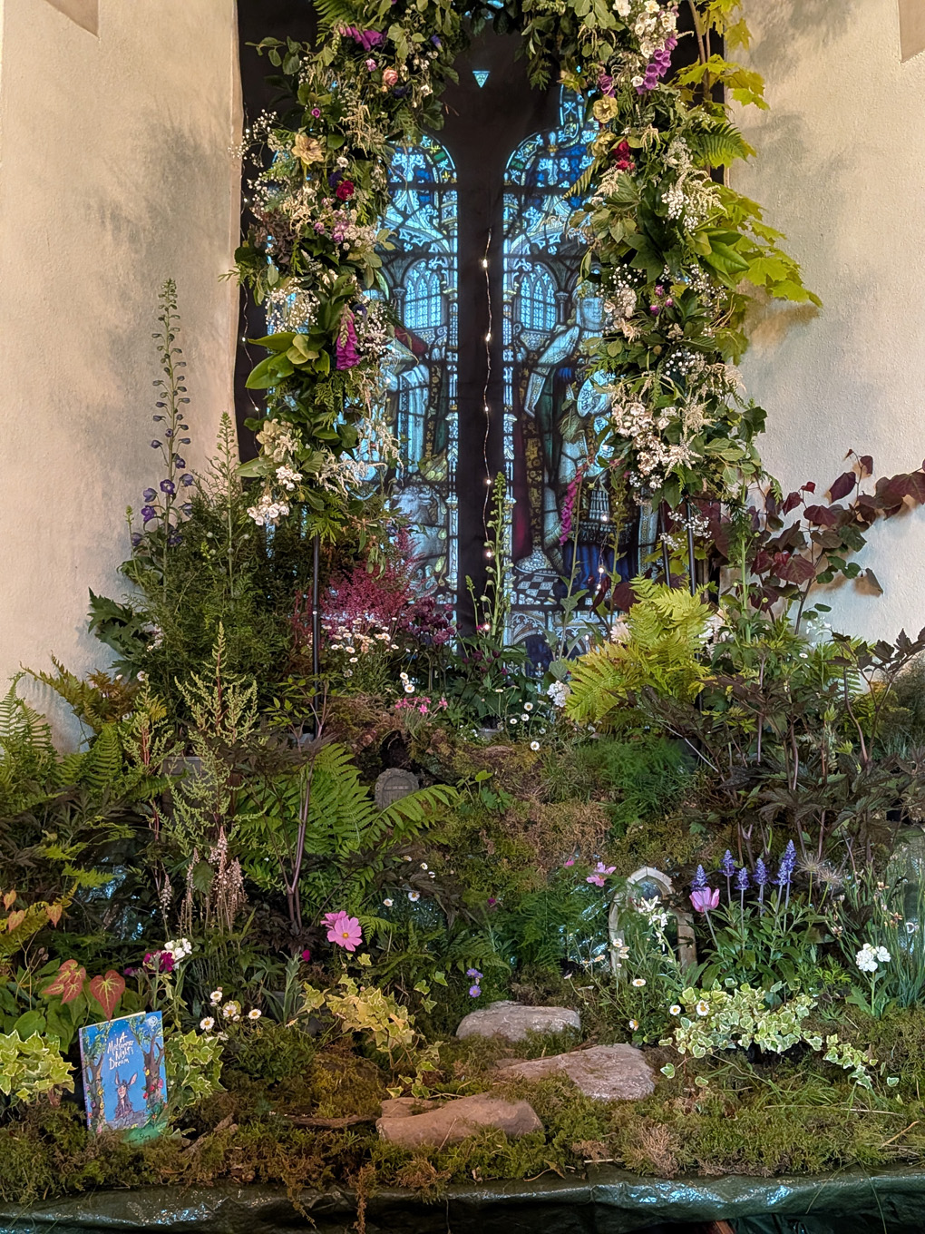 A beautiful woodland scene created with flowers and greenery around a stained glass window.