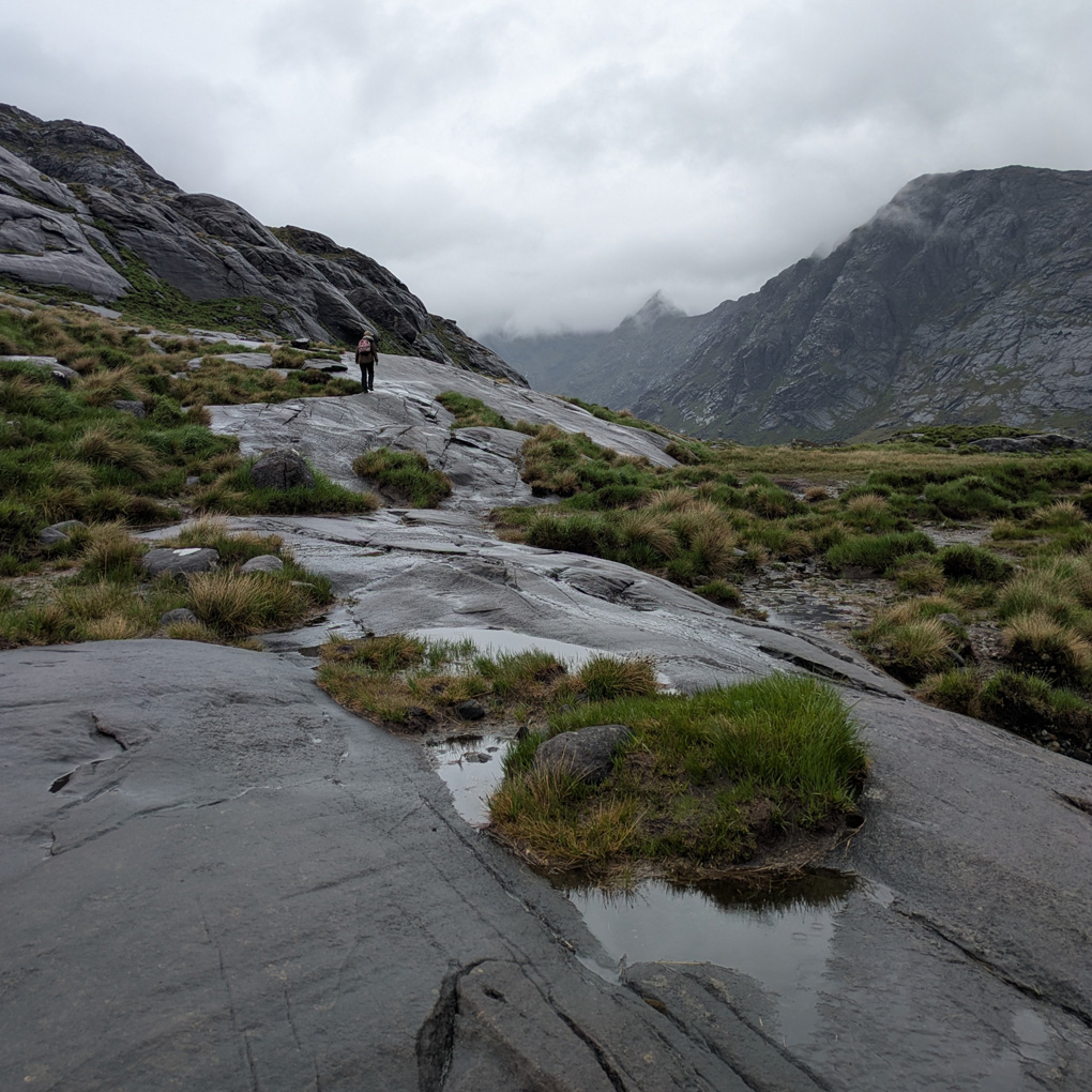 A small figure is walking ahead up on the ancient weathered rock that rises beside Loch Coruisk on grey day with low cloud enveloping the peaks of the steep hills around