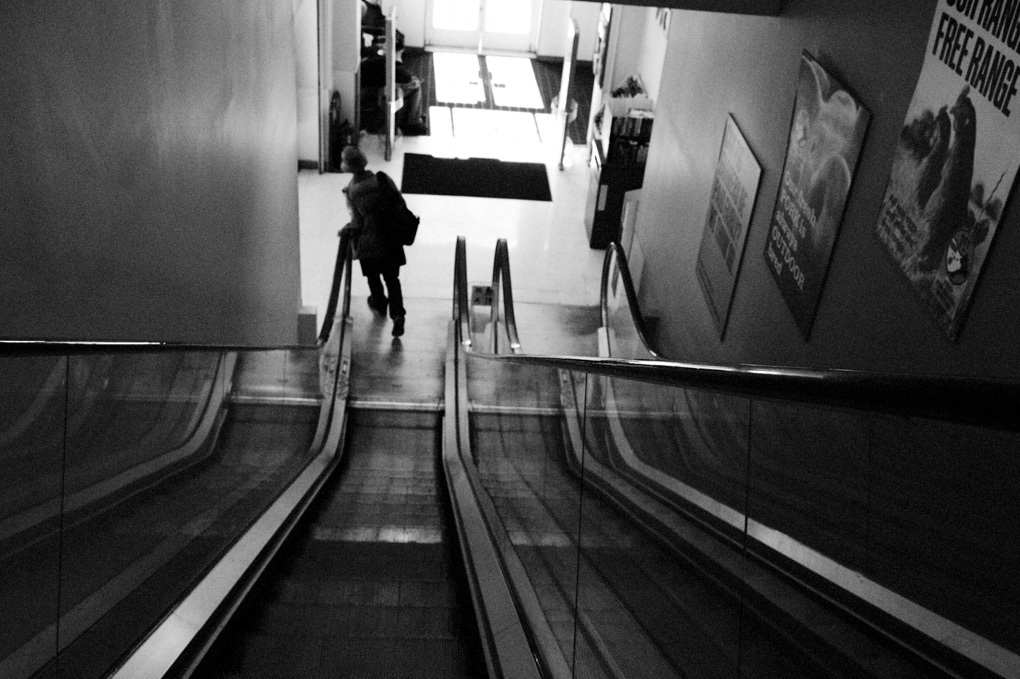 A black and white image shot from the top of the escalator in the Podium, looking down from the library landing. a man is tilting to his lift as he fast walks around the corner from the bottom of the escalator to the doors to the street. strong contrast from the black of the escalator and the man's silhouette with the light glinting off of edges in the late afternoon.