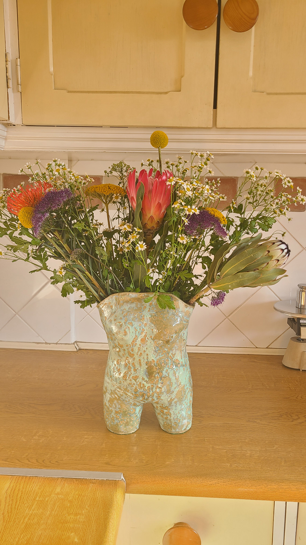 A selection of brightly coloured and interestingly textured flowers in a vase shaped like a woman's torso in a swimming costume. On a brown kitchen counter with white tiles behind.
